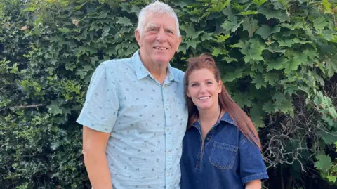 Mike and his daughter, Kate. They are standing in front of a hedge. Mike has short white hair and Kate has long auburn hair. She is wearing a blue denim dress and he is wearing a blue shirt. They have their arms around each other and are looking directly at the camera and smiling.
