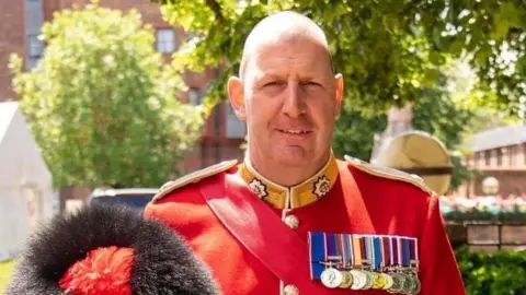 Vern Stokes is stood looking at the camera wearing a red soldier uniform, displaying his medals