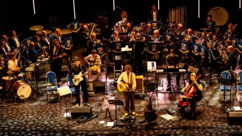Gordon Armstrong Bill Ryder-Jones sings into a microphone on stage, accompanied by his band and a crowd of men and women, who form the brass band. 