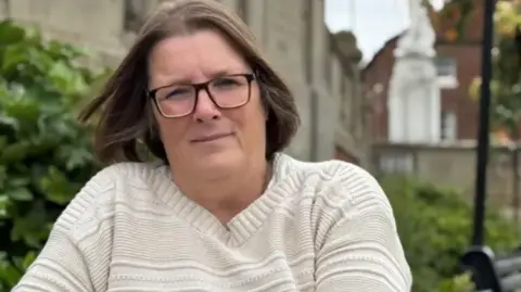 A woman with long brown hair, glasses and a white jumper with bushes behind her