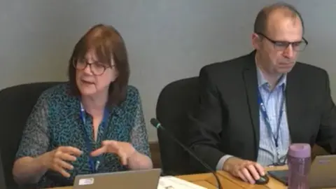 Solihull Council Beate Wagner is wearing a patterned top and has shoulder-length, brown hair. She is in a meeting room with a laptop on her desk as she addresses people and has her hands in an expressive motion