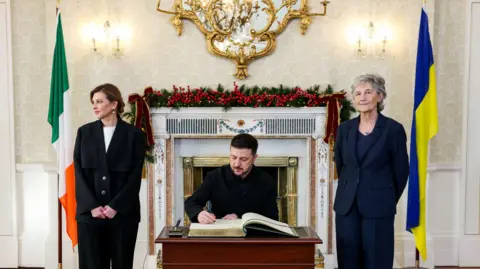 PA Media President Volodymyr Zelensky, signing the visitors book, his wife Olena Zelenska (left) and President Catherine Connolly in her study at Aras an Uachtarain in Dublin during his visit to Ireland. It is an ornately decorated room with a green carpet. A desk with a large book is in the middle.