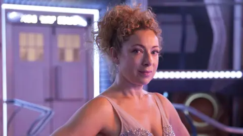 Alex Kingston has curly blonde hair and is standing in front of the exit of the Tardis, from Doctor Who.