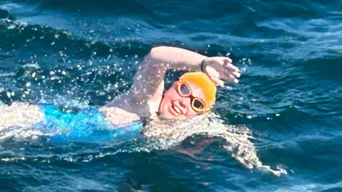 A teenage girl swimming in the sea with a blue swimming costume, orange swim cap and orange goggles on. She is mid-stroke with her right arm in the air and out of the water and is smiling towards the camera.