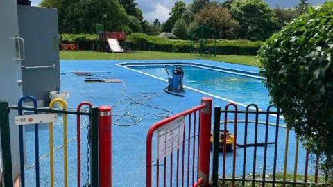 Shariqua Ahmed / BBC A blue paddling pool in the middle of the park with trees and a red entrance gate 