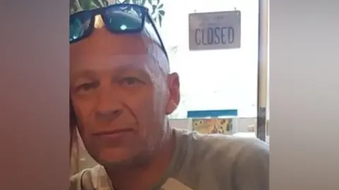 Lee Abbott is posing for the camera inside. He has sunglasses resting on his head and is wearing a grey T-shirt. A sign reading CLOSED is hanging on a glass window or door behind him.