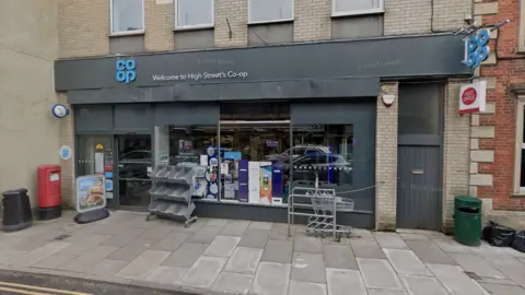 Google Google maps street view of the Co-op along Malmesbury's High Street. The building is grey with a blue Co-op logo on the top. A trolley, a red post box a newspaper stand are outside the shop. A Co-op sign and post office sign are on the side of the building. 