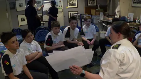 A woman in uniform shows photos to a group of young cadets