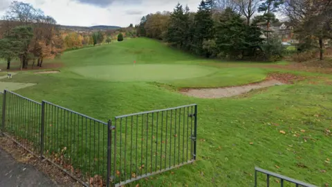 General view of Google T-shirts at Dalmuir Golf Course. There is a grey fence in the foreground of the image. On the tee, golfers in dark clothes are swaying a club. He was surrounded by green plants in the course.