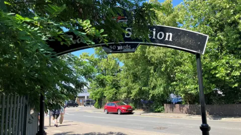 Submitted A black sign saying Rail Station. The arched sign is over a pavement. It is partially covered by leaves. A road with a single red car parked on it and more trees can be seen beyond the sign.