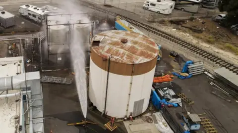 An aerial view shows a large chemical tank being sprayed with a stream of water