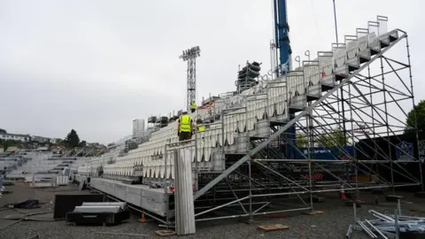 TRIVANDI Workers wearing high-vis yellow jackets constructing a stand at an outdoors sports venue.