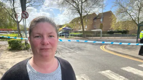 Kirsty Knight wearing a grey T-shirt and a black cardigan. She is standing in front of the crime scene. There is blue-and-white police tape in the background.