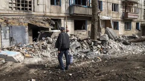 A person looks towards the rubble of an apartment block on a street in Kostiantynivka after Russian shelling on 13 March. They're wearing a cap and looking away from camera, and mud is seen on the street next to the pile of rubble with the building in the background.