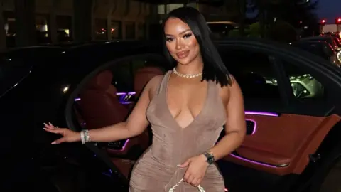 A woman in a light brown dress stands in front of a car, with a shell necklace and straight black hair.