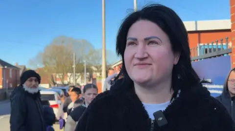 A women with short black hair wearing a white t-shirt with a black zip-up fluffy hoodie on. She is on the school run and is looking just off from the camera smiling.