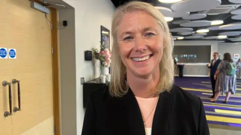 Jill Britton is wearing a black blazer and pink jumper. She has blonde hair and is stood in a hotel lobby. 