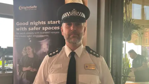 Head and shoulders of Ch Insp Andy Ainsworth, in police uniform without a jacket, standing by a poster that says "NightSafe - good nights start with safer spaces"