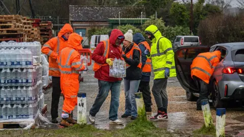 Getty Images Multiple people standing in the rain in jackets next to large pallets of bottled water. One man is carrying a case of water towards a parked vehicle.
