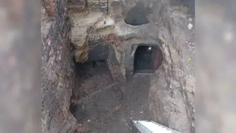 Supplied A view of a cave entrance with a few doorways and stairways visible in the right-hand background. In the foreground is a modern ladder leading into an opening before the stairways.
