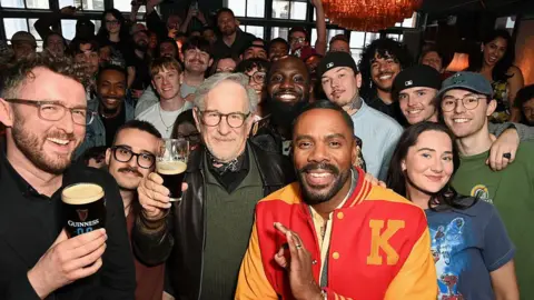 Getty Images Film director Steven Spielberg (C) in a black leather jacket, green jumper and bandana around his neck, poses with fans inside a London pub while holding an half-full glass of Guinness. To his left is actor Colman Domingo in a red and yellow letterman jacket.