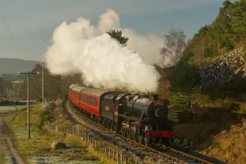 Walter Ritchie A steam train with a green carriage for the driver and several red passenger carriages, comes round the corner while billowing smoke