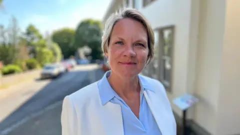 BBC Woman in her 40s with short blonde hair. She wears a white blazer and a light blue shirt