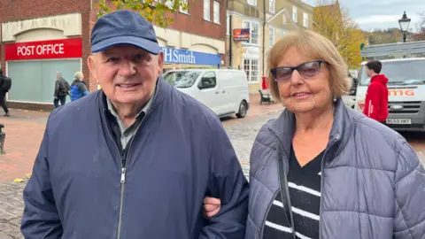 BBC / George Carden An elderly man in a navy cap, wearing a navy jacket is standing next to an elderly woman who is wearing a silver puffer jacket and a navy and white striped jumper. They are standing on a street in the middle of town.