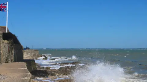 Janey Grew A British flag can be seen blowing in the wind on the left of the image and the main photograph is looking out to the sea on a bright day, with a wave sending a spray of water towards the camera.