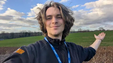 Isak Sandgren-Clarke in a selfie taken on a training walk. He is wearing a black jumper and a lanyard with 'staff' written on it. He has quite long dark blonde hair and stubble and is smiling. His arm is outstretched towards the distance.