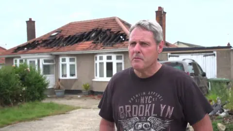 Dawid Wojtowicz/BBC Neighbour David stands in front of the fire struck home. He is wearing a dark coloured t-shirt and behind him you can see the burnt out remains of the roof.