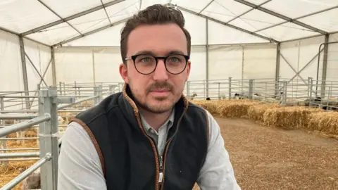 Kate Bradbrook/BBC Jack Pishhorn with very short dark beard and glasses sitting on a straw bale in a marquee