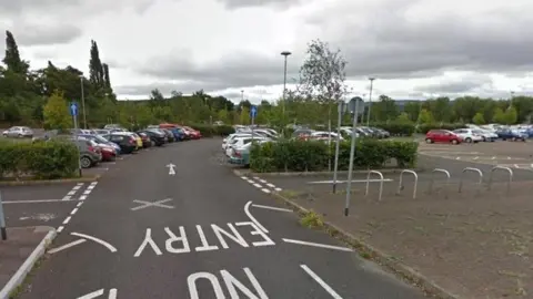 Google A wide view of the Taunton Gateway park-and-ride site showing an entrance road and parked cars. The car park area is bordered with trees and bushes