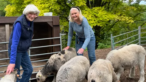 Two women smile widely while surrounded by sheep. Behind them is a small copse of trees