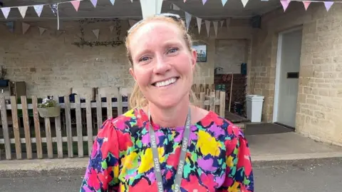 Chris Wood/ BBC A woman smiles for the camera. She has tied black blond hair and is wearing a flower-print dress. In the back ground is a limestone wall with some potted flowers attached