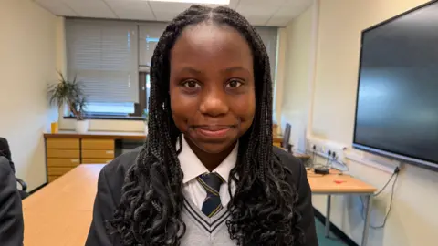 Kytenda pictured with long dark braided hair smiling at the camera with closed lips. She is wearing a white shirt with a black blazer and navy striped tie.