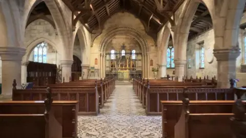 Inside St Edith's church there is a tiled floor consisting of an intricate mosaic design which stretches all around the building. Several lines of wooden pews line the way to the altar which includes brass figures. Light from the stained glass windows seeps through, illuminating the edges of the church. 