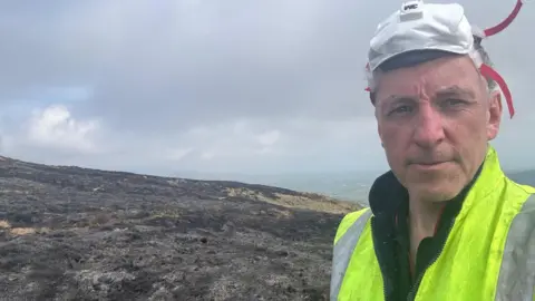 Vincent McAlinden Vincent McAlinden who has short grey hair and has a mask pushed up on top of his head stands on a charred hilltop