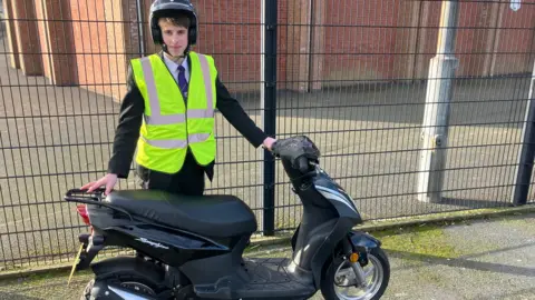 A male pupil at St Mark's High in Warrenpoint. He is wearing a helmet, has brown hair and wearing his school uniform -  black blazer, white shirt blue tie, and black trousers. He is standing behind a black moped.