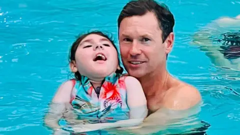 Family photo Darcey, a brunette haired girl wearing a blue and red swimming jacket with her father Stephen, who has short dark hair, in a swimming pool