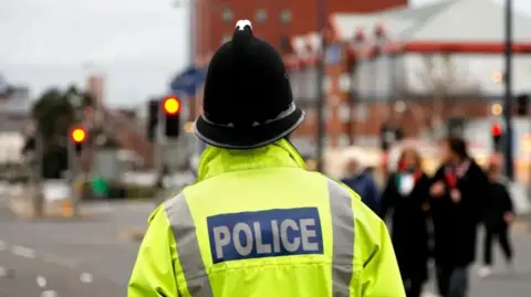 File photo of a police officer, standing with his/her back to the camera and wearing a police helmet and yellow hi-vis jacket with POLICE on the back.
He is standing next to a traffic junction in a busy town or city. Pedestrianised can be seen crossing the road behind him.