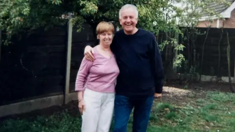 Stephen Hindley Judith and Steve Hindley standing together in a garden, with Judith wearing a light pink top and pale trousers and Steve in a dark jumper and jeans, his arm around Judith, in front of a wooden fence and leafy trees.
