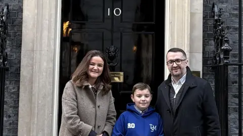 Contributed Roman stands in the middle of his dad Ryan and MP Jenny Riddell-Carpenter outside 10 Downing Street. Roman wears a blue hoodie, has short dark hair and holds a letter written on a piece of paper. Ryan has short dark hair, glasses and wears a black coat. Jenny has long dark hair and wears a long grey coat.