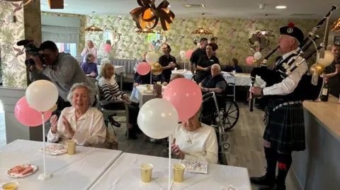 BBC/TomWalker A piper dressed in a kilt plays the pipes in a community room while elderly care home residents clap their hands while sitting at tables decorated with birthday decorations. 