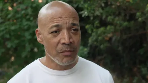 Matthew Phillip in a white t-shirt in front of greenery.