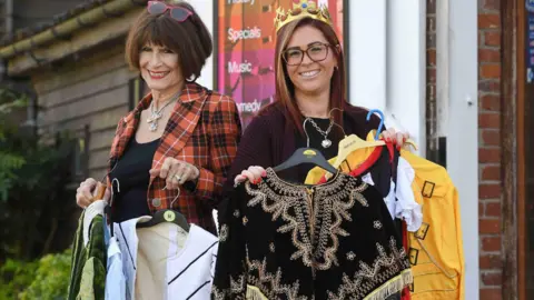 Quay Theatre, Sudbury Julie on the left wears a red and black striped jacket and lakc top. She has a large necklace and sunglasses on her head. She is holding coat hangers containing a variety of costumes. Krystle is holding other costumes and wearing a crown on her head. They are standing outside the theatre