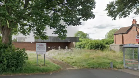 A methodist church set back from the pavement. The red brick building which has three white crosses in front of it is surrounded by overgrown grass and bushes. There is a bus stop on the far right and a church sign on the left. 