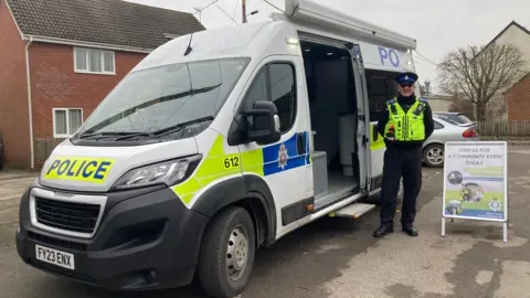 A large police van with an officer standing outside smiling at the camera. Parked in a residential area, it has a sign outside, inviting people in for commmunity engagement.