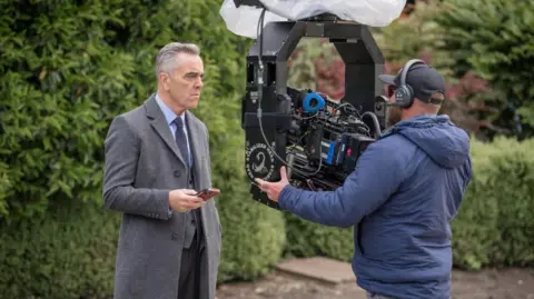 James Nesbitt, who is wearing a grey coat with suit and tie, is standing in front of a man who is holding a camera in a park during filming. 