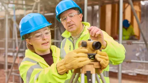 A female construction worker stands behind a builder's level on a building site. Behind her a co-worker walks across the development.